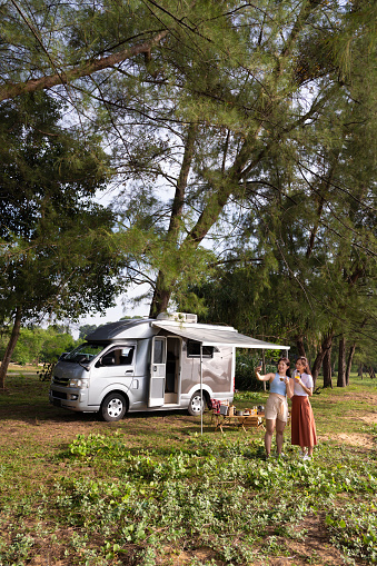 Two Asian women taking selfie photo together in front of camper vehicle during a caravan picnic holiday vacation. Family Day, Travel & Tourism, Summer Holiday Vacations, Travel with Camper Van Concepts.