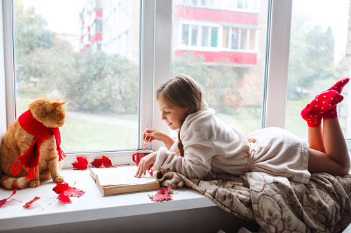 Little girl with red cat lying on window sill , reading a book and drinking cocoa. Winter weekend with cat at home.