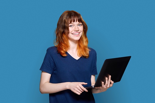 Red-haired teenage girl with laptop on blue background. Adorable smiling teenage female student looking at camera. Youth, technology, education leisure lifestyle concept