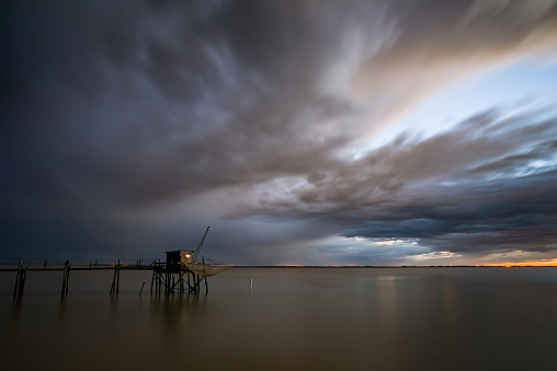 Dark thunderstorm clouds at sunset over fishing hut and pier standing in flat calm water of Gironde estuary, Charente-Maritime, France near La Rochelle
