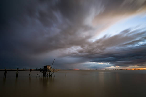 Stormy weather sunset over calm seas Dark thunderstorm clouds at sunset over fishing hut and pier standing in flat calm water of Gironde estuary, Charente-Maritime, France near La Rochelle calm before the storm stock pictures, royalty-free photos & images