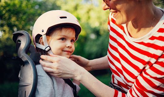 Close up photo of woman hands fasten the helmet of excited little girl sitting in bike seat of parents.