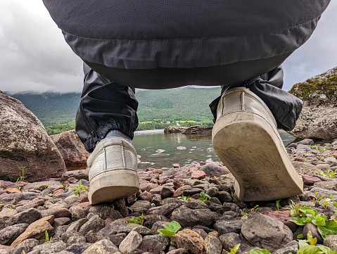 Close-up view between the feet of a crouching hiker on a water edge of Norway fjords, resting on a pebble rocks beach looking at the valley