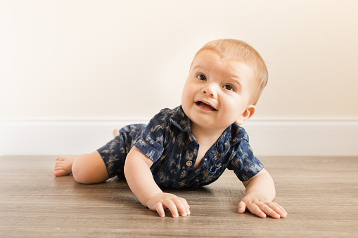 7-Month-Old Baby Boy With 12 Toes Laying on His Belly On a Wooden Floor While Wearing a Blue Linen Button-down Romper.