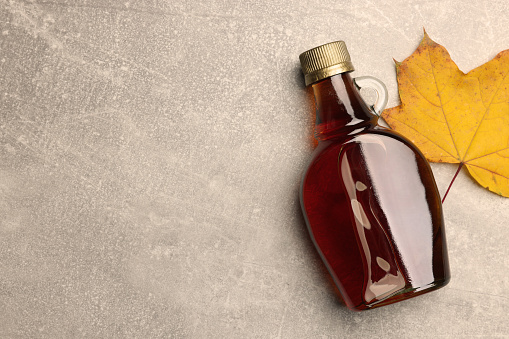 Bottle of tasty maple syrup and dry leaf on light grey table, flat lay. Space for text