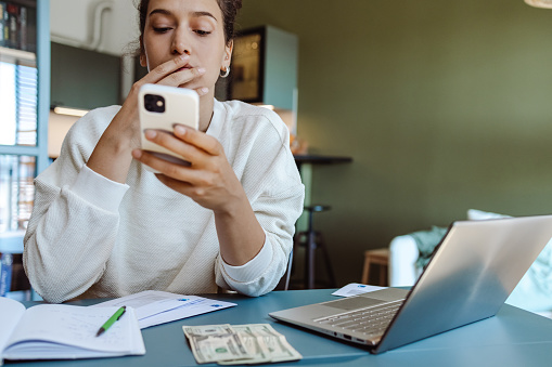 Young woman in a beige sweater sitting at the table and calculating expenses and taking care of personal finances. She is using smart phone while calculating personal expenses