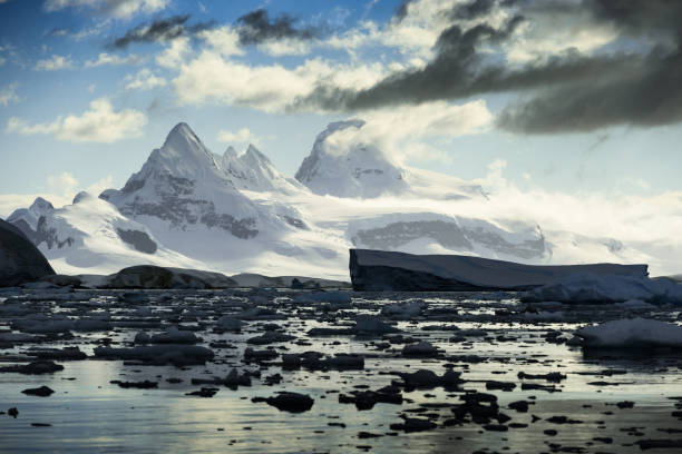 Being at water surface level makes the Antarctic views all the more dramatic. In the shadows of the bay, the sun glistens on the distant mountains.  Frigid water surface reflects the clouds of the sky.