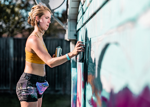 Female artist painting large wall mural she designed. She is dressed in casual work clothes with shorts and sport top bra. Exterior of large wall of the bungalow house.
