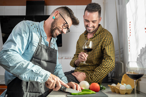 Happy hipster gay couple in love hugging while standing in the kitchen. They are cooking dinner together at home and drinking wine. High quality photography.