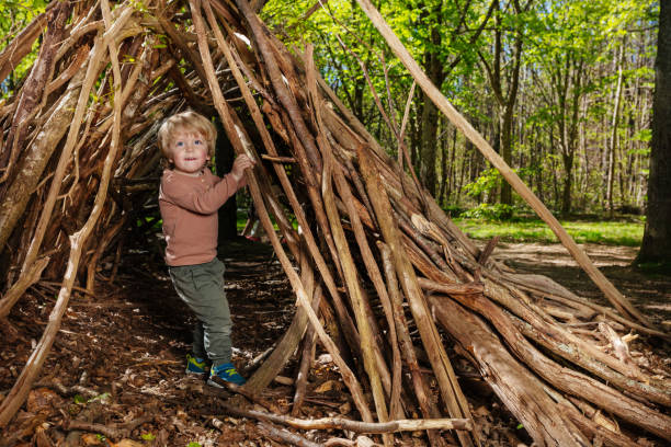 Little blond boy play in hut of branches in the forest, happy time in summer camp