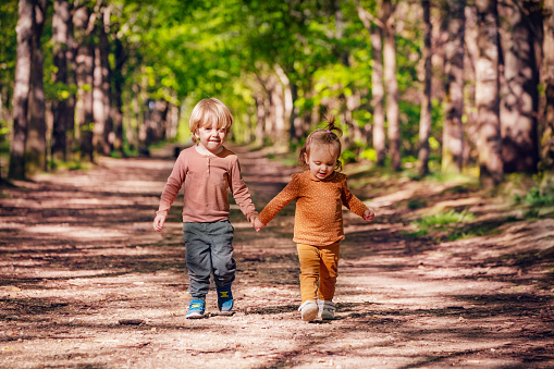 Two little kids blond boy and a girl walk holding hands on the alley in the park at summer, playing catch-up