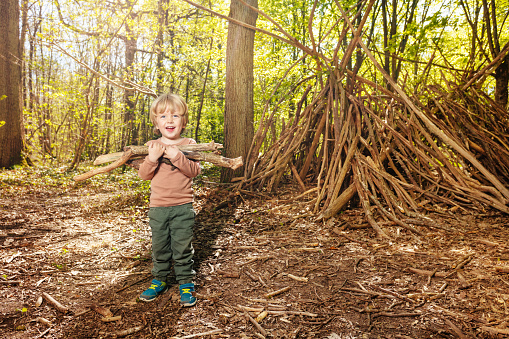 Boy with pile of brushwood in the forest build hut of branches, happy and laughing