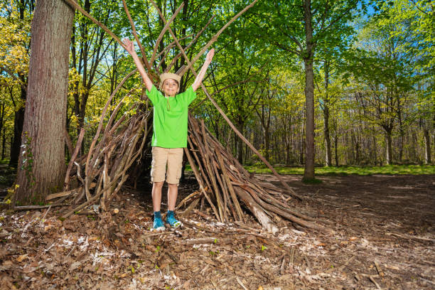 Preteen young boy in boy-scout hat plain in the forest hut of branches lifting hands up and smile in summer camp