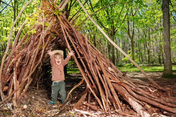 Little blond boy in boy-scout hat plain in the forest hut of branches lifting hands up and smile at summer camp