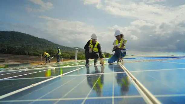 Team contractor, male engineer and female technicians  wearing safety uniform, talking about installing plan, check the working system and maintenance solar panel of solar power plant to produce electricity on the roof of factory building. Industrial of renewable energy. Power generation from solar energy.