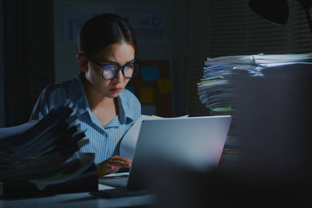 empleado de oficina asiático con exceso de trabajo que usa una computadora portátil para trabajar a altas horas de la noche en la oficina. trabajar horas extras por la noche - effort dark concepts women fotografías e imágenes de stock