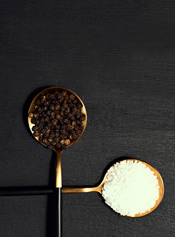 Salt and pepper on spoons against a black wooden background. Seasalt and peppercorns seasoning for cooking a flavorful or tasty meal. A table with condiments to add flavor to food