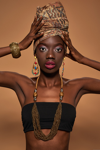 Partial of stylish black girl wear traditional african outfit and accessories look at camera. Young woman wearing tank top, turban, necklace, earrings and bracelet. Orange background. Studio shoot