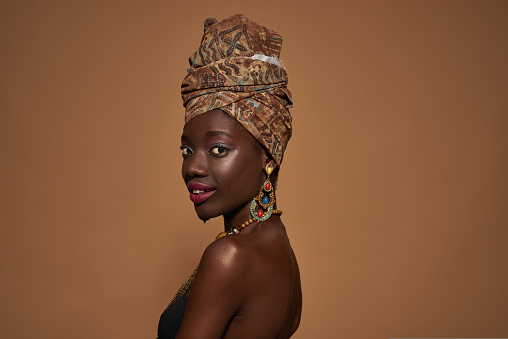 Side view of smiling fashionable black girl wearing traditional african outfit and accessories looking at camera. Pretty young slim woman. Isolated on orange background. Studio shoot. Copy space
