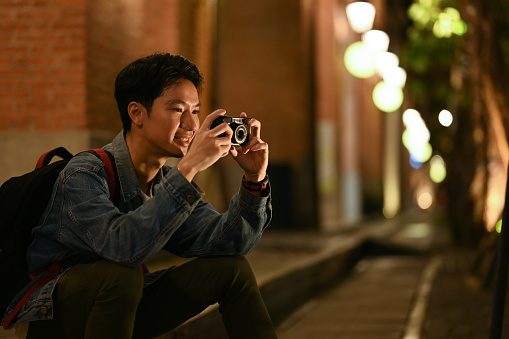 Young male traveler taking photo with his camera, sitting on stairs in the night city with blurred night street lights background.