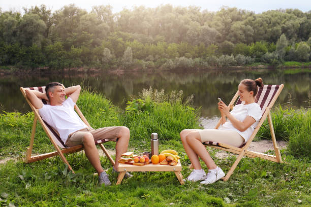 Outdoor shot of young couple sitting on deck chair and talking near the river, relaxed man sits with hands behind head, woman using cell phone for checking social networks, family relaxing outdoor. foto al aire libre de una pareja joven sentada en la tumbona y hablando cerca del río, un hombre relajado sentado con las manos detrás de la cabeza, una mujer que usa el teléfono celular para revisar las redes sociales, una familia que se relaja al aire