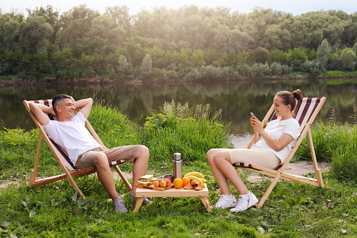 Outdoor shot of young couple sitting on deck chair and talking near the river, relaxed man sits with hands behind head, woman using cell phone for checking social networks, family relaxing outdoor.