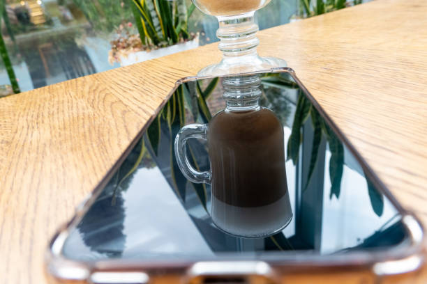 latte with foam in transparent glass on wooden table in cafeteria. There are plants indoors. Silhouette of glass is visible on black screen of mobile smart phone, lying on table. Mirroring effect. Cozy relax atmosphere in restaurant.