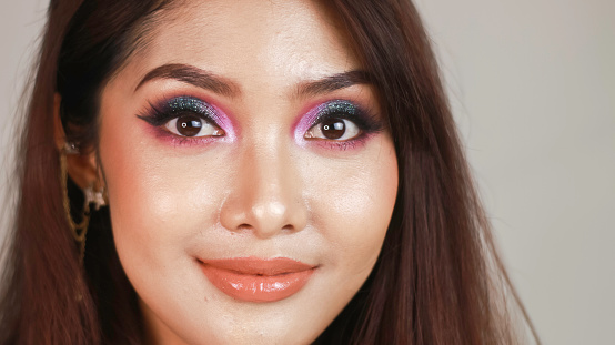 A beautiful young Asian girl with colorful make-up on a white background looks into the frame.