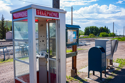 A telephone box that once had a paid telephone, is rewired with a standard telephone that is set in rural scene near railroad tracks along a dirt road on a sunny day.