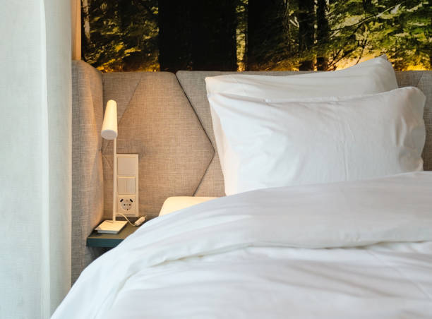 istock Fragment of bedroom with bedside table, reading lamp and socket in modern interior​ design home or hotel. Soft pillows and blanket, stylish comfortable furniture. Dark wall and room, selective focus. 1413970885