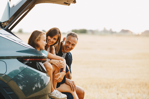 Pretty couple in sweaters and hats sitting in the car trunk on the forest roadside. Young family traveling by car in the pine forest