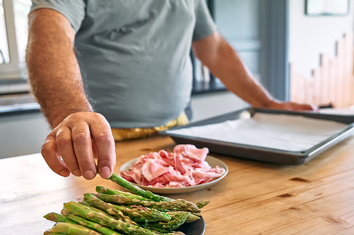 Man preparing young green asparagus sprouts wrapped in bacon on wooden table in the kitchen. Man's hand taking asparagus from plate.