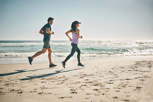 Fit, active and running couple on beach for workout, training and exercise by sea or ocean with copy space. Sporty, athletic and healthy man and woman jogging for cardio health, stamina and endurance