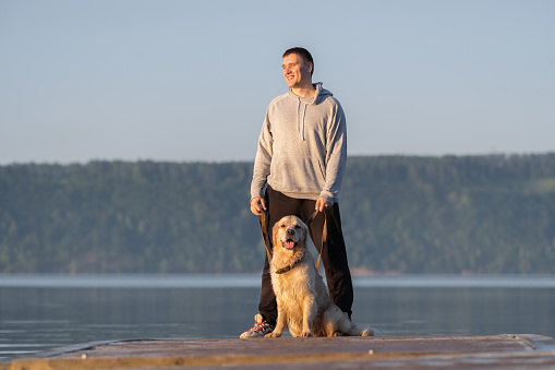 A man and his dog are sitting on the dock at sunrise in the summer. A golden retriever and his owner are walking by the river.
