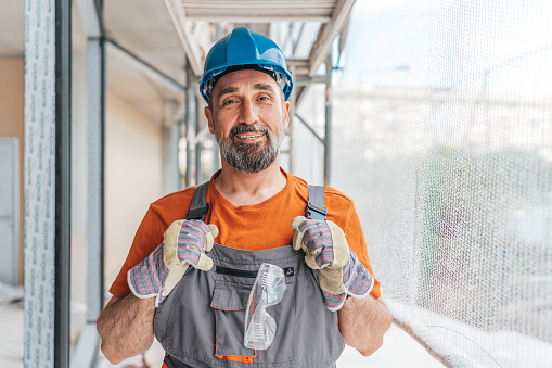 A mature Caucasian male construction worker is standing in a room under renovation and looking at the camera with a warm smile on his face.