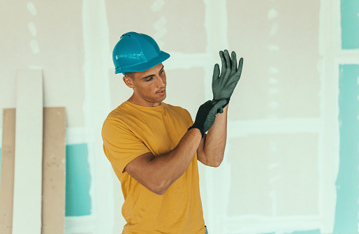 A young Caucasian male construction worker is standing in front of a wall under renovation and putting on safety gloves.