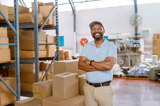 Portrait of a smiling worker, arms crossed in a warehouse in the delivery business. Man in storage distribution centre with packaged parcels. Logistics employee in a factory storehouse with box goods