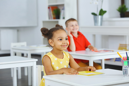 A Black girl in a yellow dress listens attentively to the teacher. She holds a copybook in her hand