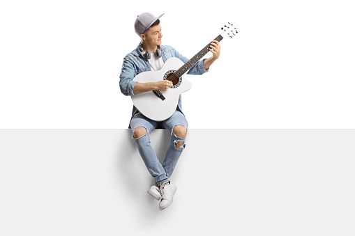 Guy sitting on a blank panel and playing an acoustic guitar isolated on white background