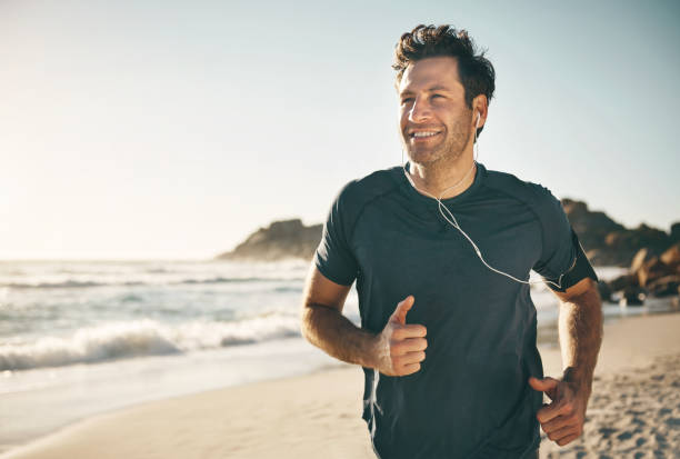 Active, fit and healthy man jogging on a beach while listening to music on earphones with stunning outdoor clear sky and sea copy space. Happy, mature and athletic guy running, doing fitness exercise stock photo