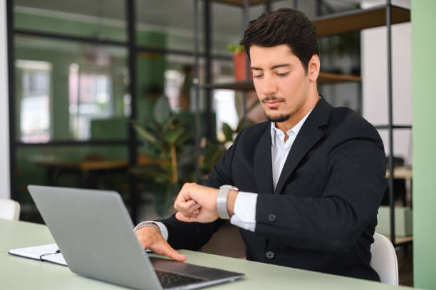 Busy young bearded entrepreneur or male employee in formal wear sitting in office Busy young latin male entrepreneur or employee in formal wear sitting in office and working on laptop, checking the time from a watch on his wrist, time to start online conference or virtual meeting Time Management  for Entrepreneurs stock pictures, royalty-free photos & images