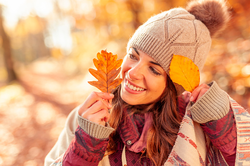 Cheerful young woman having fun spending sunny autumn day in nature, holding colorful fallen leaves and peeking behind them