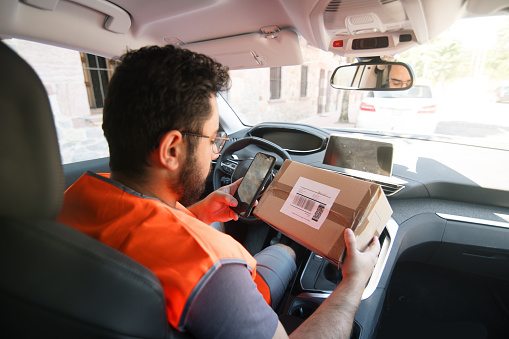 A young delivery man scanning a parcel tag with a smart phone inside a car before giving it to the destinataire