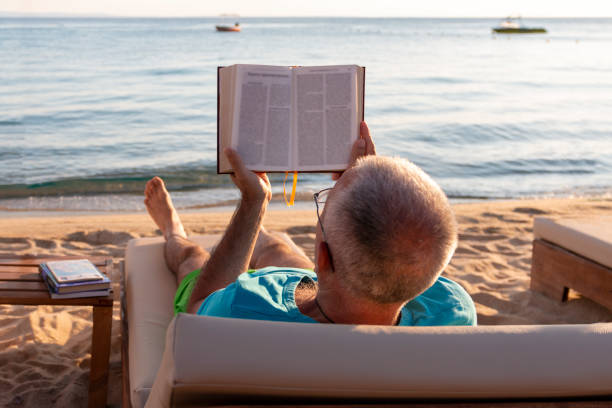 istock Mature man lying on a sunbed and reading a book at sunset. 1413993595