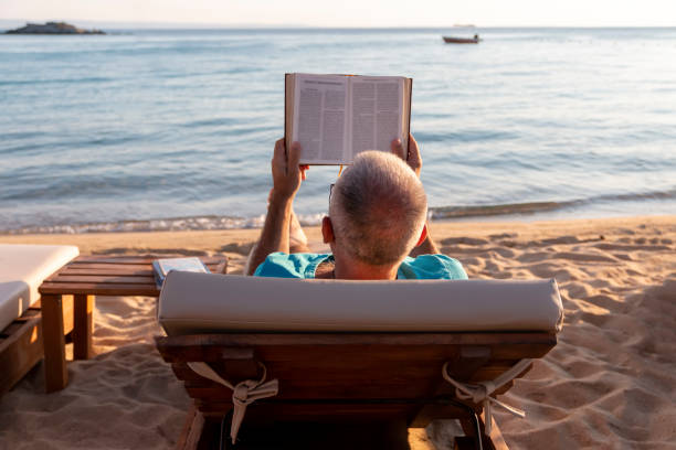 istock Mature man lying on a sunbed and reading a book at sunset. 1413993598