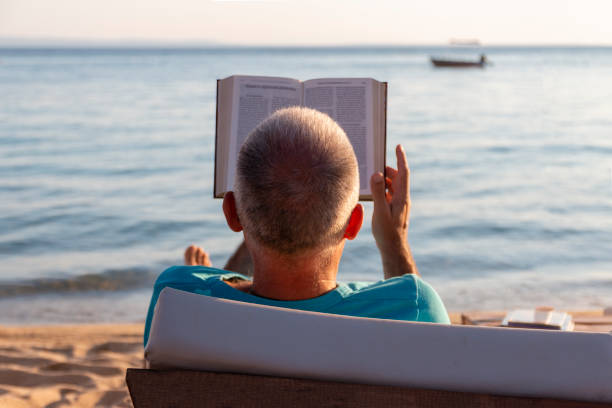 istock Mature man lying on a sunbed and reading a book at sunset. 1413993600