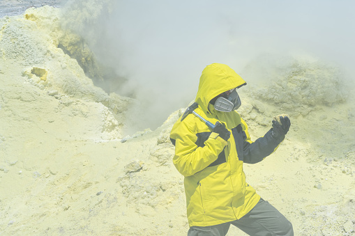 male volcanologist with a geological hammer and in a respirator on the slope of a volcano nearby among toxic fumes examines a sample of a mineral