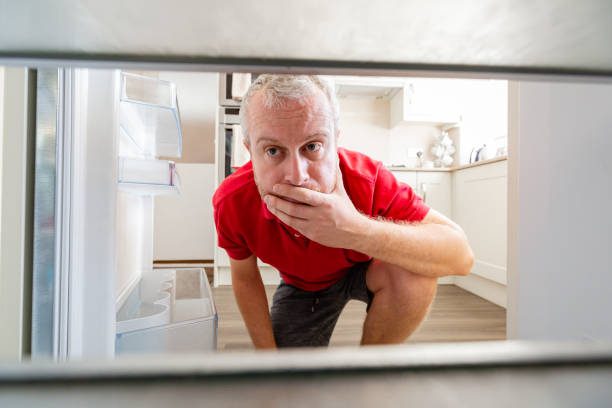 Hungry man looking inside empty refrigerator Wide angle close up depicting a mid adult man peering inside his bare refrigerator in his kitchen at home. The man has a worried, desperate expression due to the extreme lack of food on the shelves. fridge problem stock pictures, royalty-free photos & images