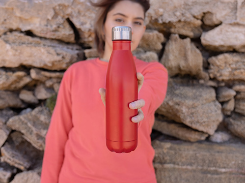 Serious Teenager in pink hoodie with red water bottle in hand looking stright. Short-haired teen girl staying outdoors near stone wall. Clothing and reusable water bottle mockup. Hipster and adolescence concept