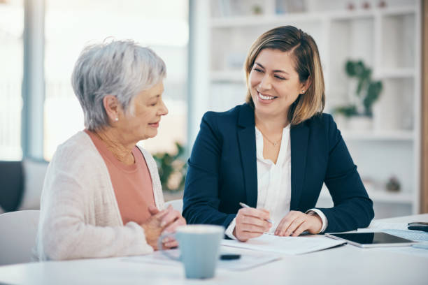A mature financial advisor consulting and planning contracts with a senior client. An old woman meeting her banker to discuss and plan her retirement finances or savings and sign contracts at home stock photo
